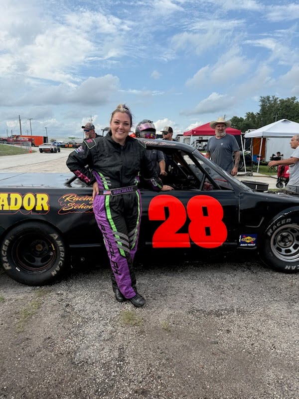 Rachelle Rudolph is in a racing suit, standing next to a race car with the number 28 at an outdoor event.